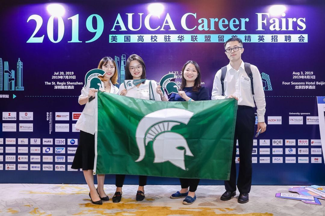 spartan students and alumni smile holding spartan flag in front of AUCA banner