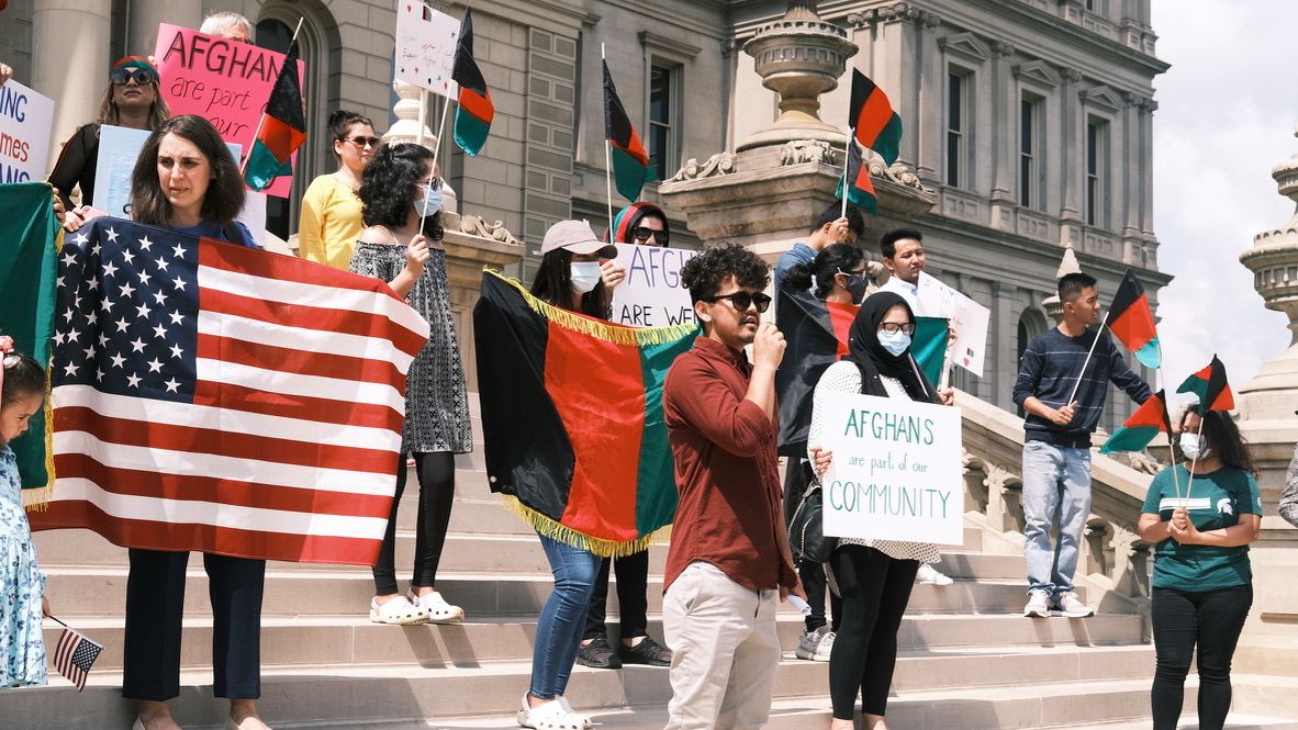 A group of people on the Michigan capitol steps holding flags and signs, one person is at the front with a megaphone