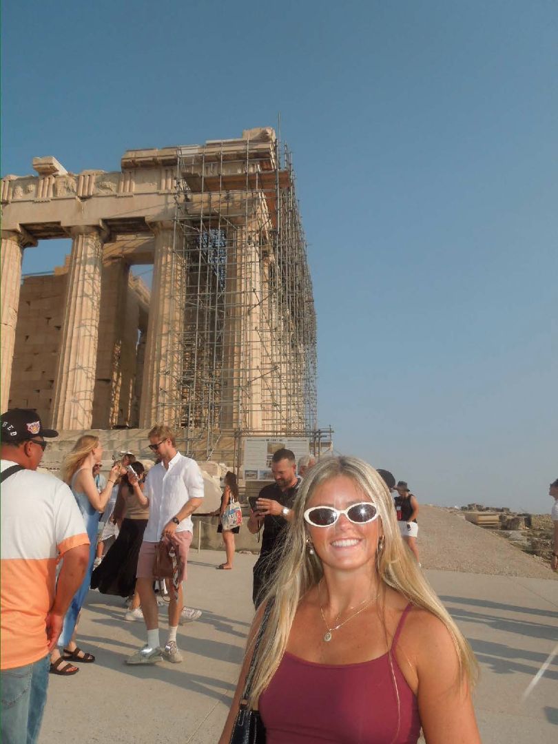 Olivia wearing white sunglasses standing in front of the Acropolis and Parthenon in Athens