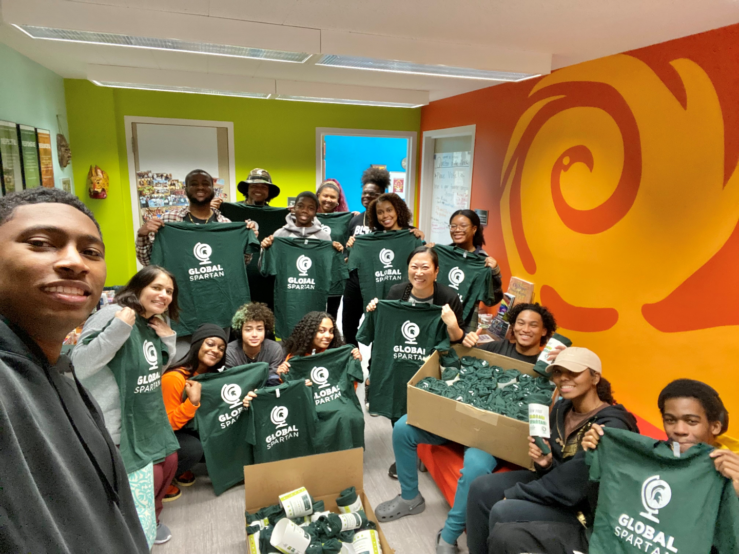 A group of students pose together holding up green “Global Spartan” t-shirts. The room has green, orange and yellow walls with a swirl design on the right one.