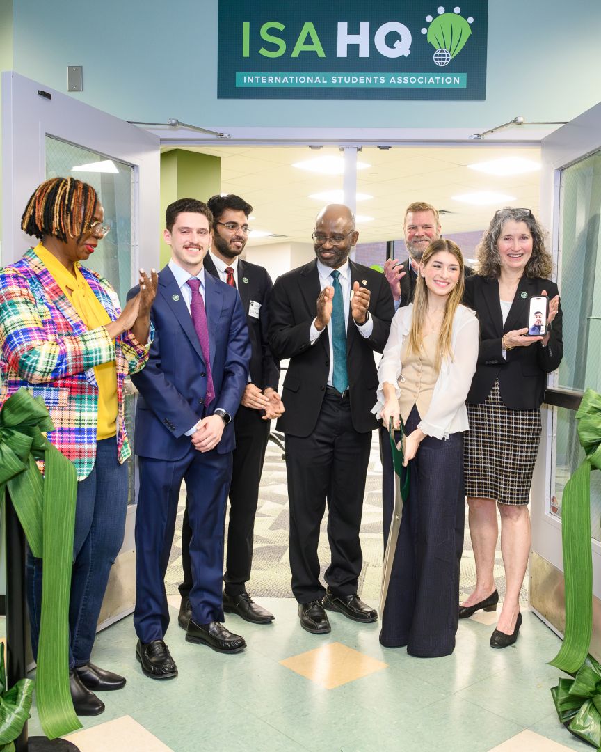 A group of people gather for a ribbon-cutting ceremony at the entrance of the International Students Association Headquarters (ISA HQ). A young woman holds large green scissors, smiling as others applaud. The diverse group includes students and university staff, standing beneath a sign that reads 'ISA HQ – International Students Association.'