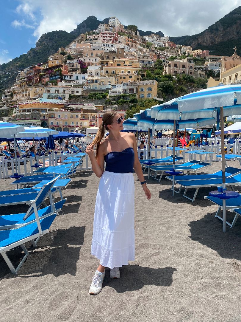 Madelynn posing on a a beach with blue lounge chair and umbrellas with colorful buidlings on a mountain in the background