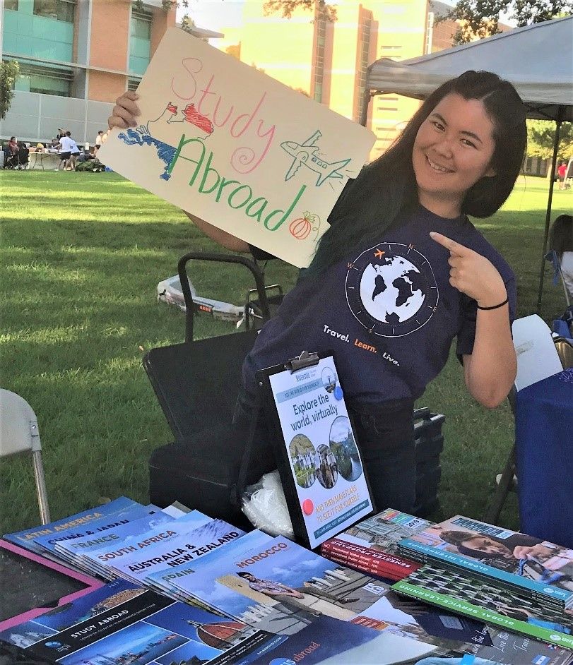 Student standing slightly tilted to the side while point with one hand to a sign they are holding up in the other hand. The sign reads "study abroad".