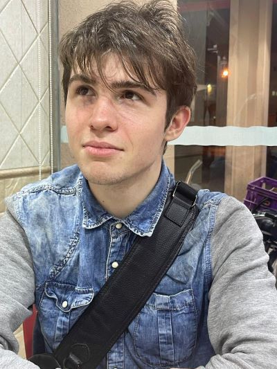 A young man sits and looks past the camera with a bag strap across his chest.