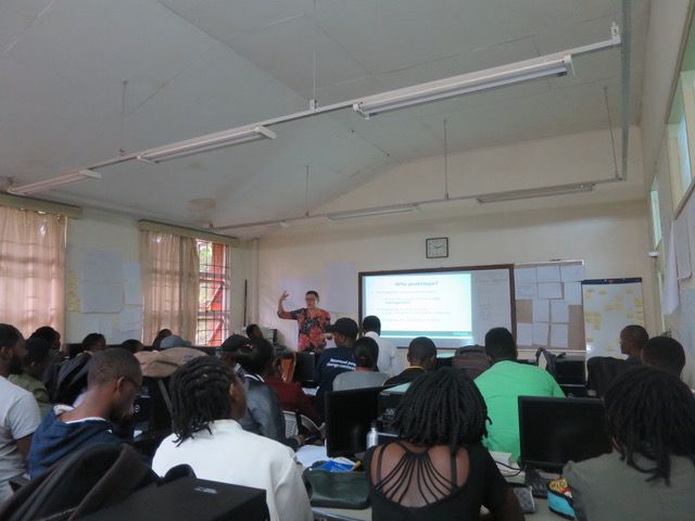 People in a classroom watch the lecturer at front speak in front of a projection screen