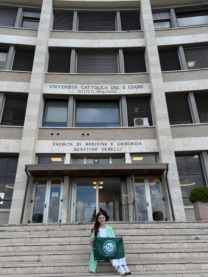 Bella sitting on the steps to the biology building of a local university hold a FLI flag