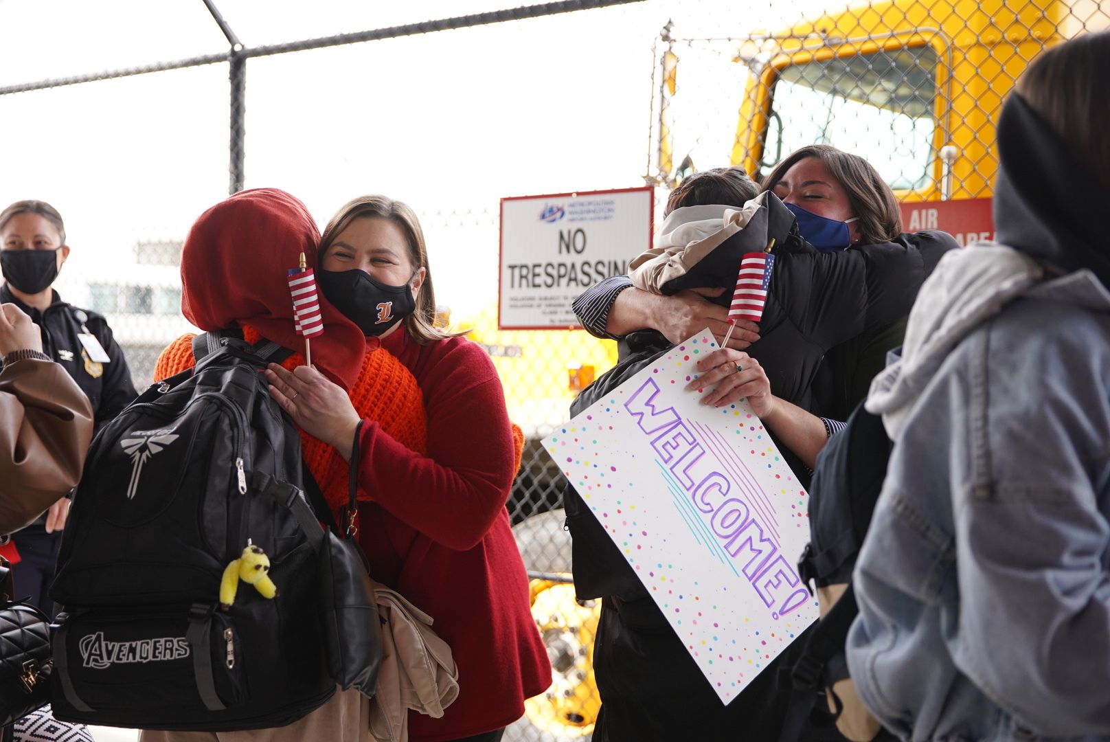 Two groups of people embrace outside of an airport. They hold an American flag and a "welcome" sign.