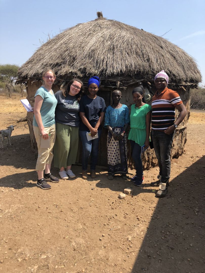 Group of students in front of hut with straw roof with local Tanzanians