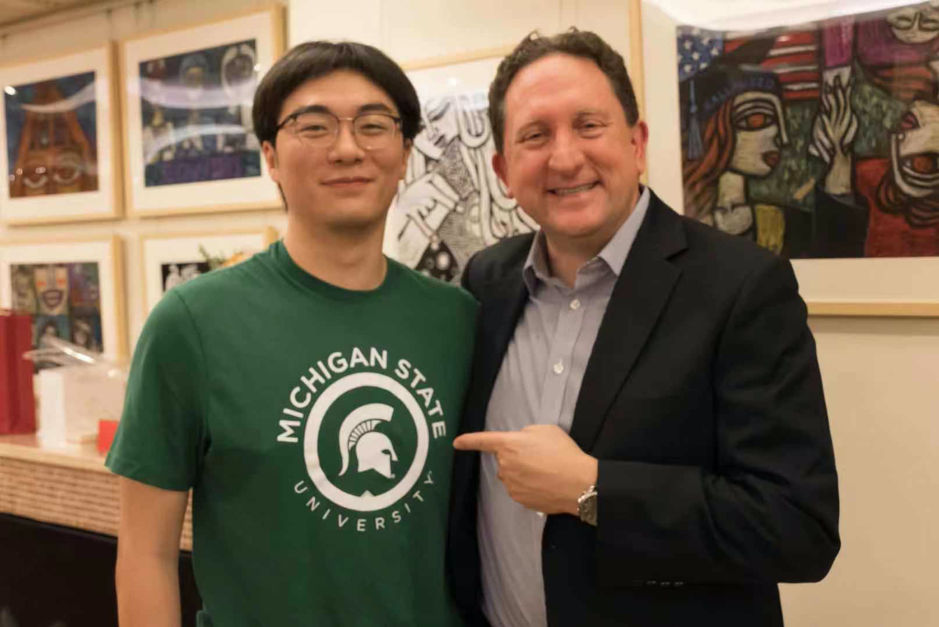 Zhicheng Hai stands beside Mr. Heller. Heller points at the Spartan helmet on Hai's MSU green t-shirt. Both are smiling.