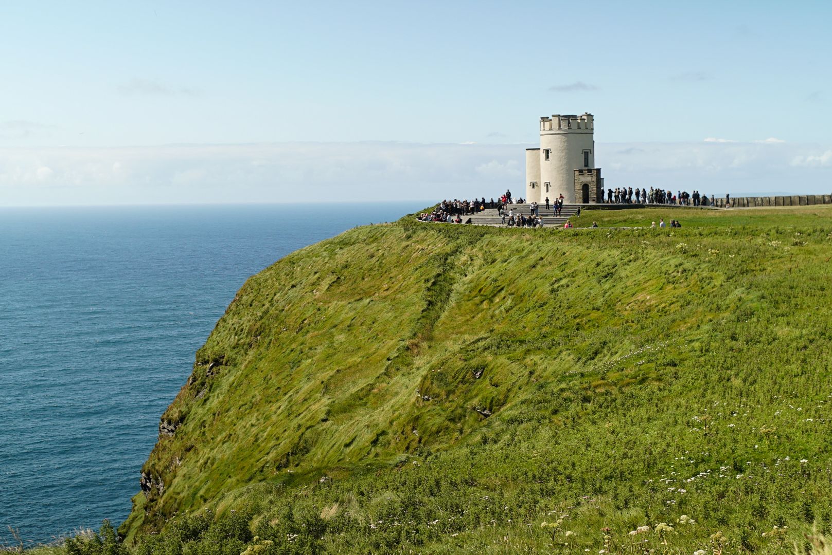 a Castle stands on top of a cliff facing the Ocean with many visitors looking out into the horizon