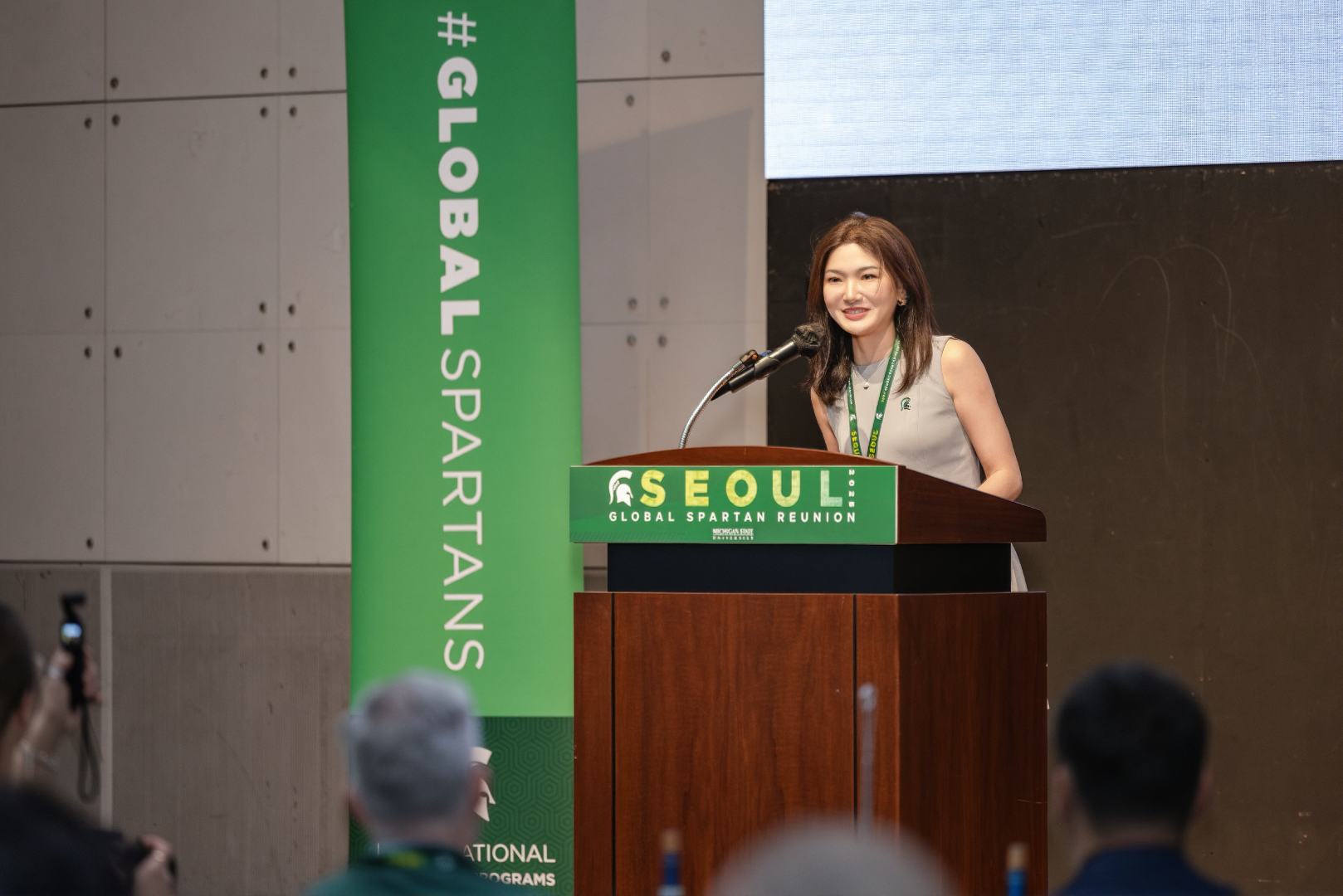 A woman stands at a podium, smiling as she addresses an audience during the Global Spartan Reunion in Seoul. She wears a gray sleeveless dress and a green lanyard. The podium is branded with a green sign reading 'SEOUL Global Spartan Reunion, Michigan State University.' A large vertical banner next to her reads '#GLOBALSPARTANS.' Attendees in the foreground appear to be listening and capturing the moment with their phones.