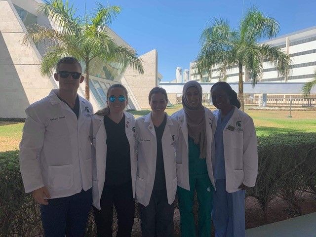Five medical students smile for the camera, wearing white lab coats in front of white buildings and palm trees.