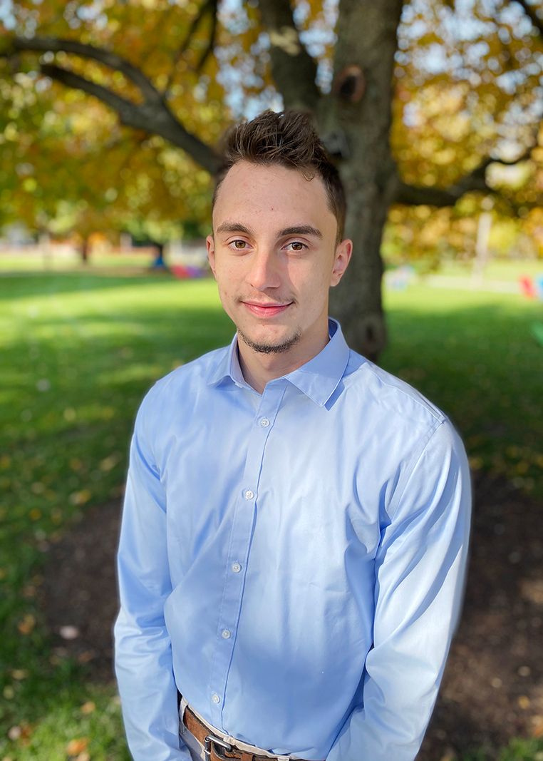 Portrait of Adam Tucek smiling at the camera, standing outside in front of a tree in the sunshine.