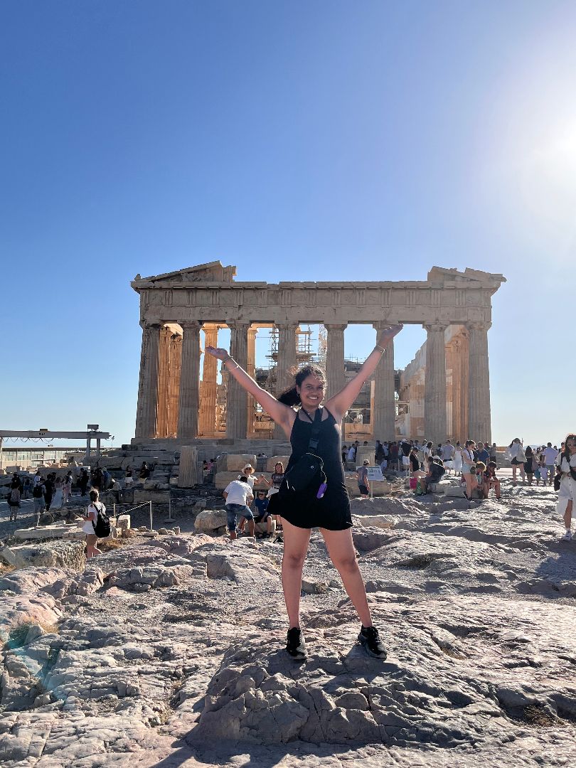 Shreya with her arm up wide standing in front of the Acroplis in Athens, Greece