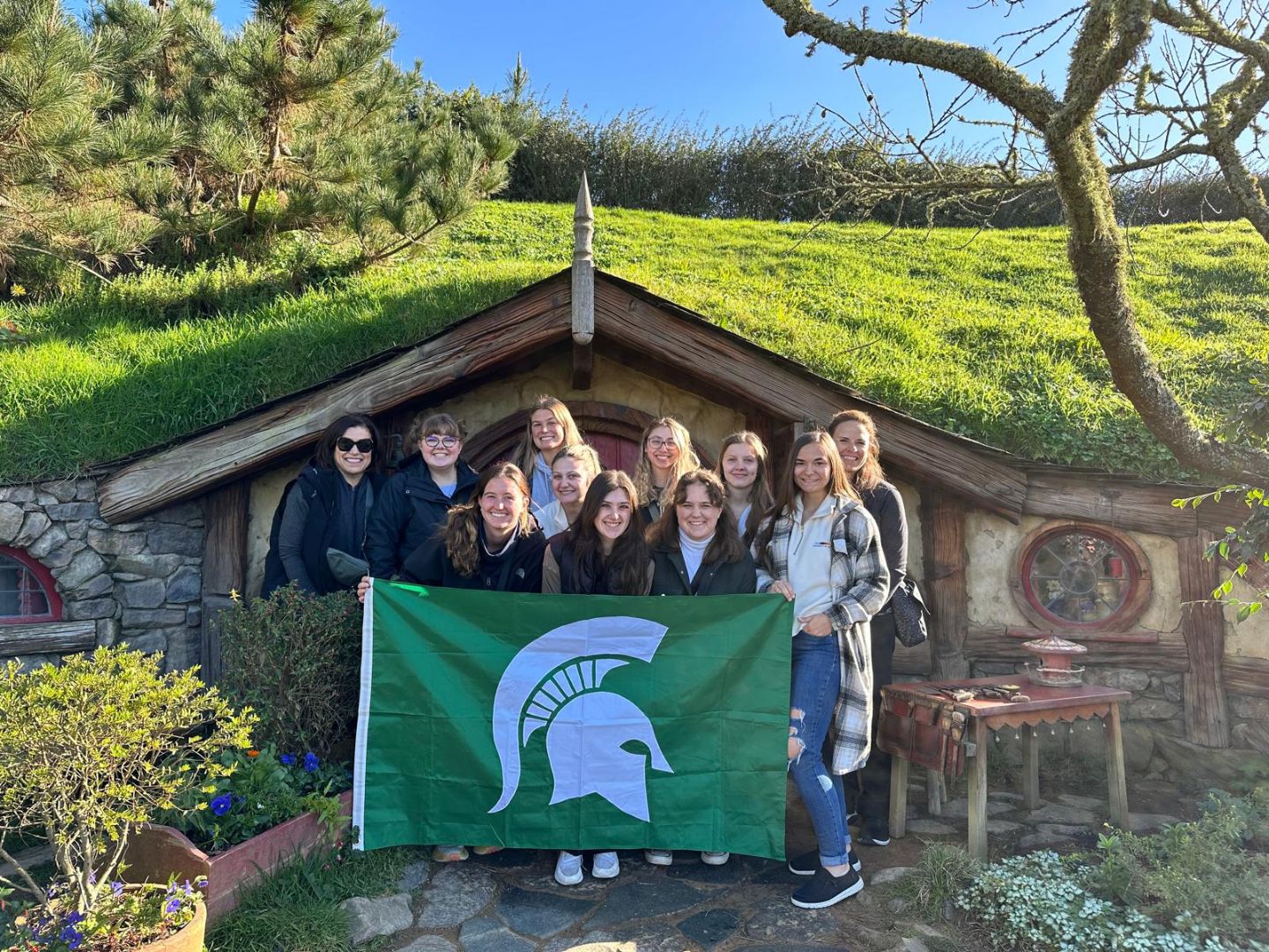 Group of students holding a Spartan flag in front of a building in Hobbiton