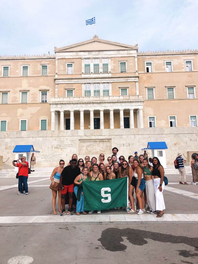 Students holding Spartan flag in front of large historic building in Greece