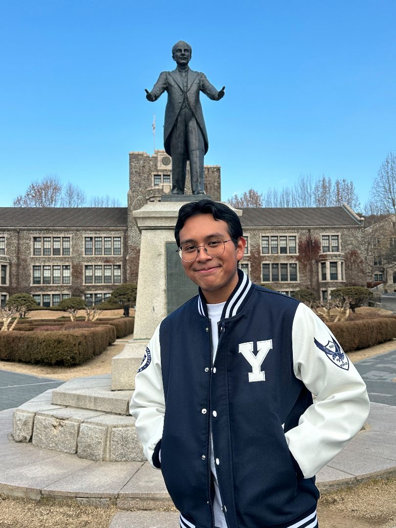 Erik wearing a blue and while Yonei letterman's jacket in front of a statue on Yonsei campus