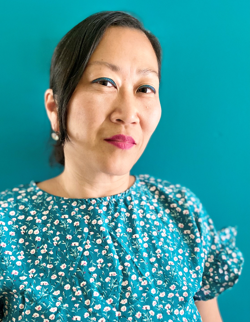 Close up headshot of a smiling woman against a teal background; she has long black hair pulled back and a blue and white floral shirt. 