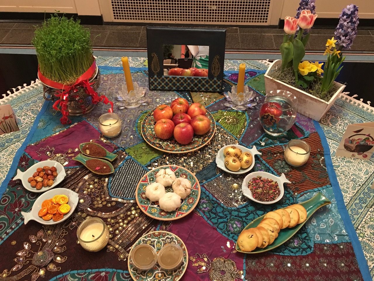 Photo of a colorful, festive Haft-Sin, set for Nowruz. There are candles, fruits, flowers, and small dishes of various foods set on top of multi-colored patchwork fabric. At the back is a framed mirror and a raised, decorative pot filled with green grass.