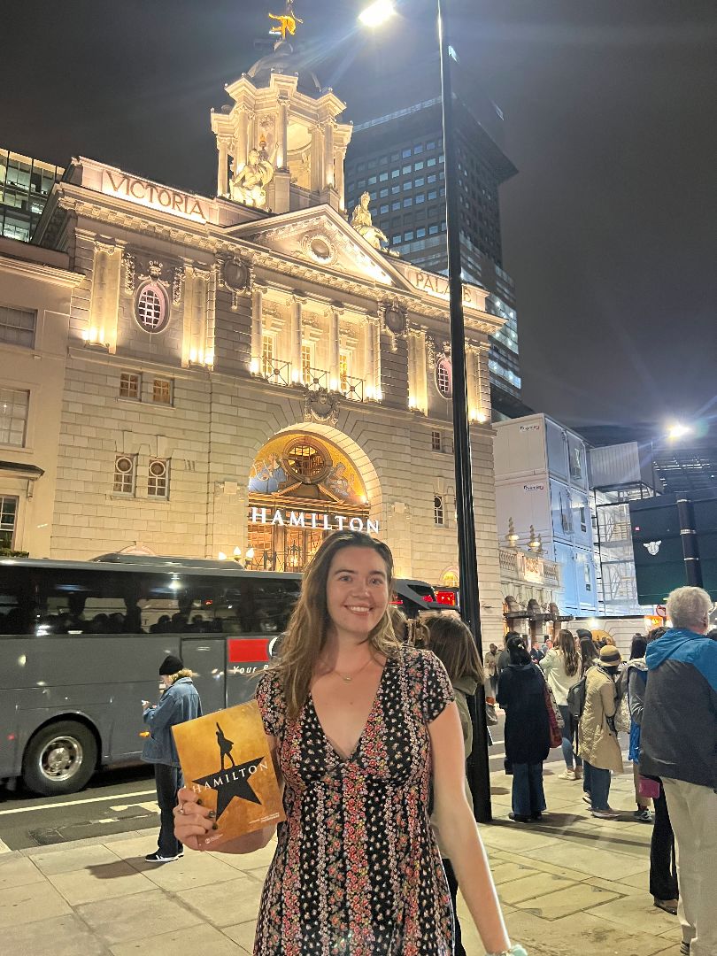 Haley standing in front of the Victoria Palace Theatre at night holding a Hamilton playbill