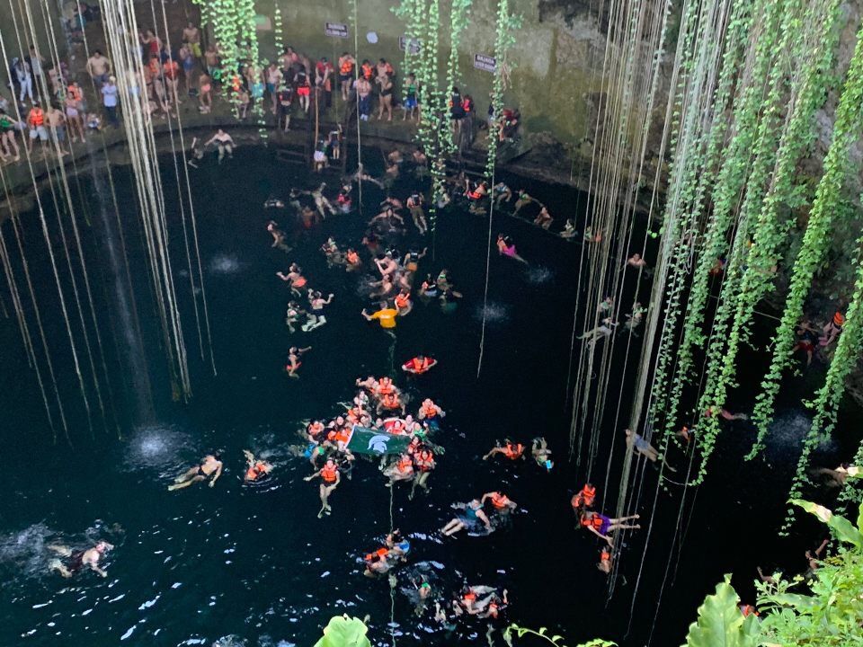 People swim in a large swimming hole in the rock, with green vines hanging down. The MSU flag can be seen in the middle of the swimmers.