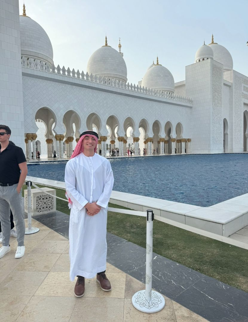 Eleftherios wearing a red and white traditional headdress and white robe standing in front of a relecting pond in Dubai