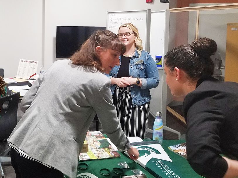 EducationUSA advisers talking to admissions counselors from MSU learning about what MSU has to offer international students. Three women standing around a table with MSU material.