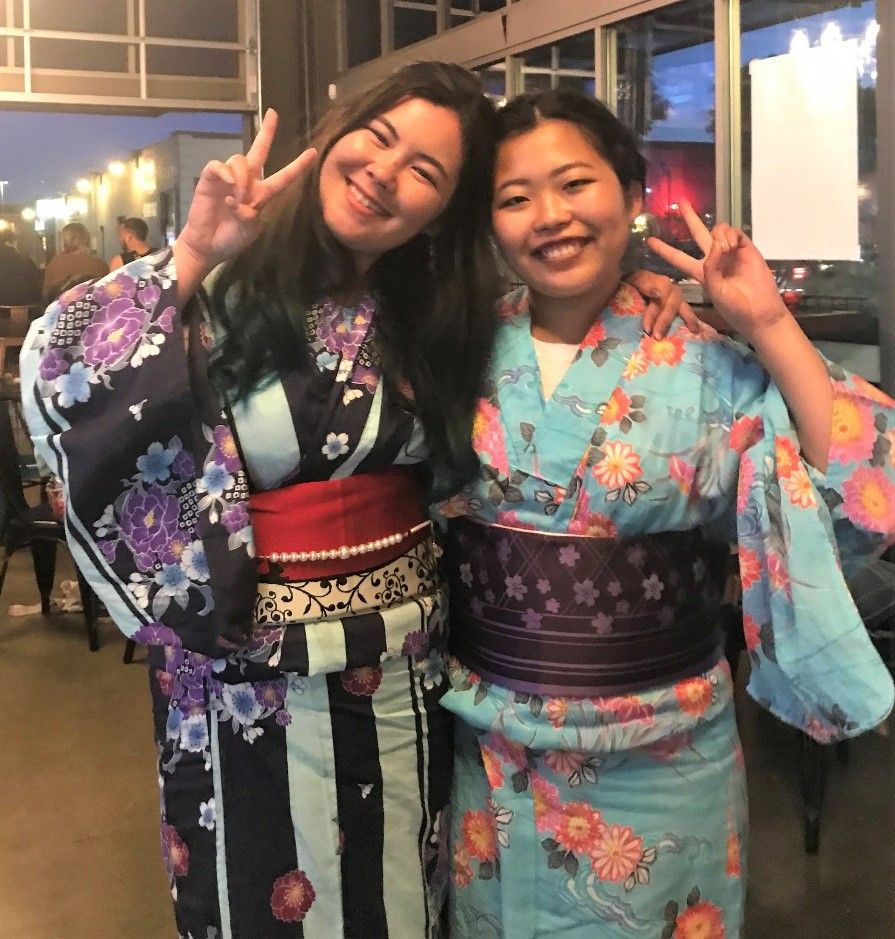 Two people standing side-by-side in traditional Japanese garb. Both are smiling and holding their hands up while making a v-shaped sign with their fingers.