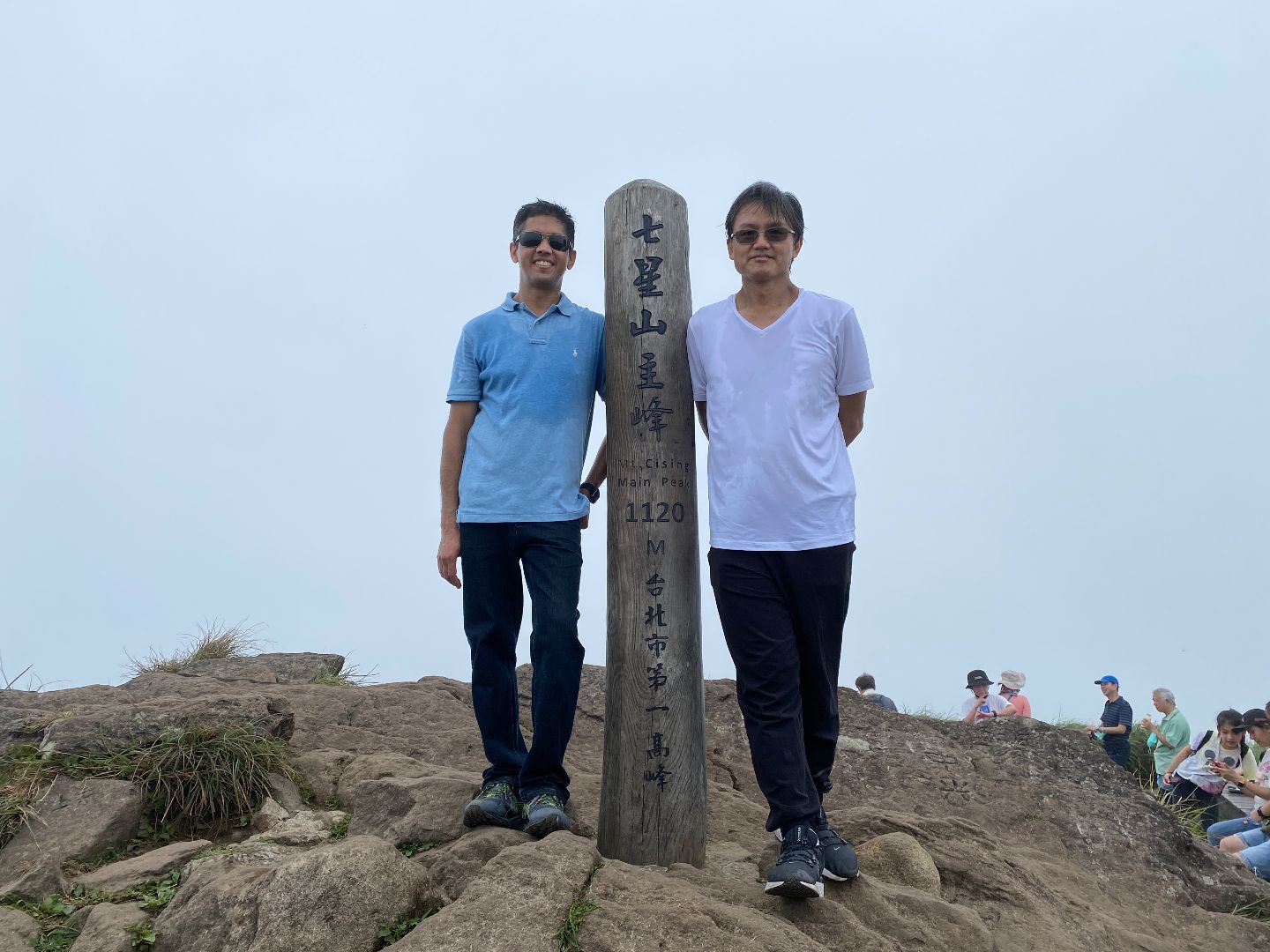 Two men stand on top of a mountain, smiling at the camera with the blue sky behind them. Between them is a tall guide post with writing on it. They are both wearing sunglasses, t-shirts and denim jeans.