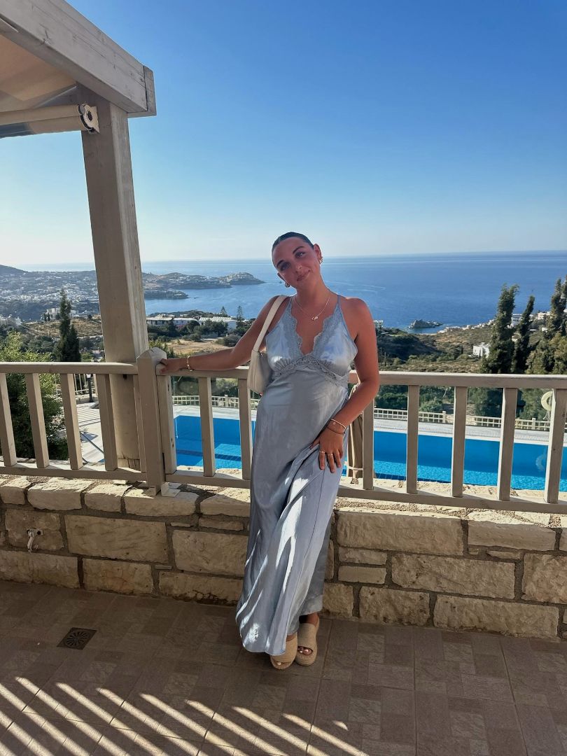 Madison in a long blue dress standing on a balcony overlooking the sea in Greece