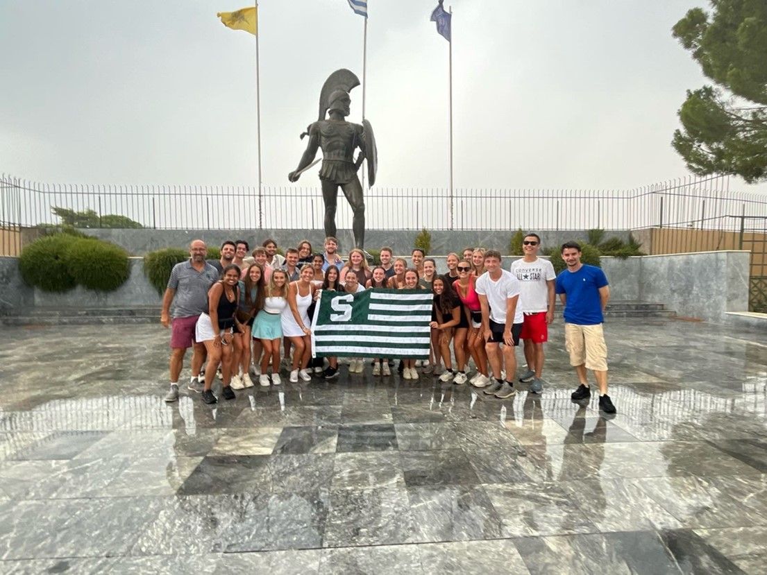 Group of students holding the Spartan flag in front of a Spartan statue in Greece