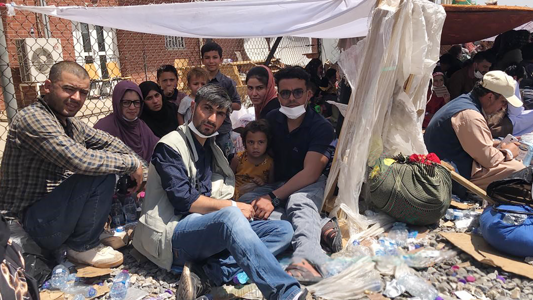 An all-ages group of people sit on the dusty ground with a makeshift tent over them