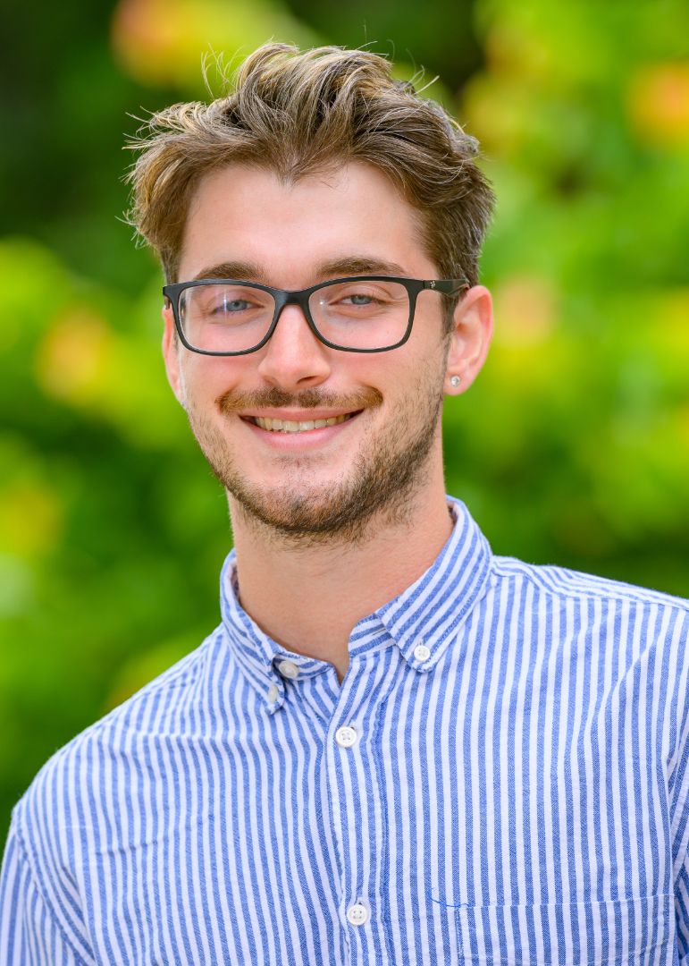 A man with short brown hair, black glasses, and a blue and white striped shirt, facing forward and smiling.