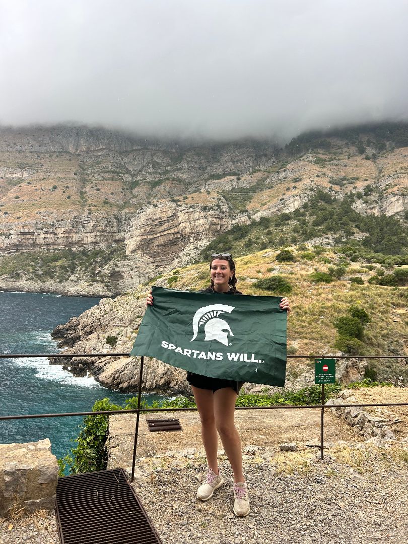 Madeline holding a Spartans Will flag by the sea in Italy