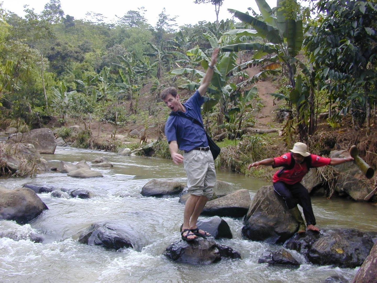 A person balances on a rock in a stream about to fall, with a colleague looking on.
