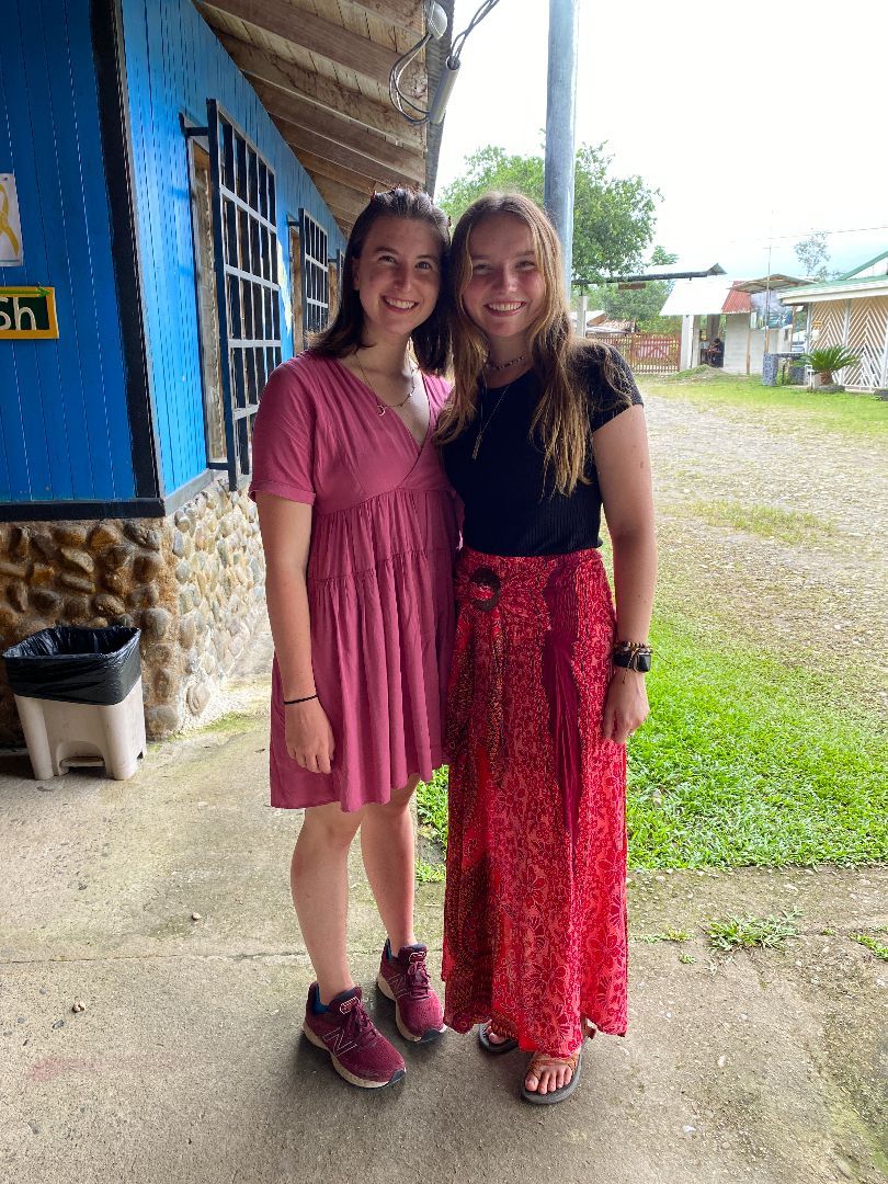 Two women stand next to one another outside smiling, under the awning of a stone building with blue siding. The woman on the left has mid-length brown hair and a flowing pink dress on. The woman on the right has long blond hair and is wearing a black t-shirt and long red skirt. 