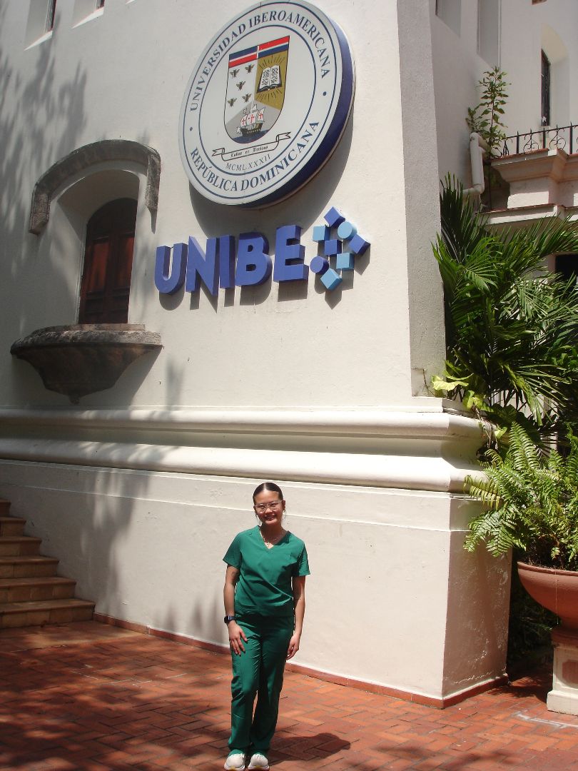 Kaitlynn in green scrubs standing in front of a building that says UNIBE