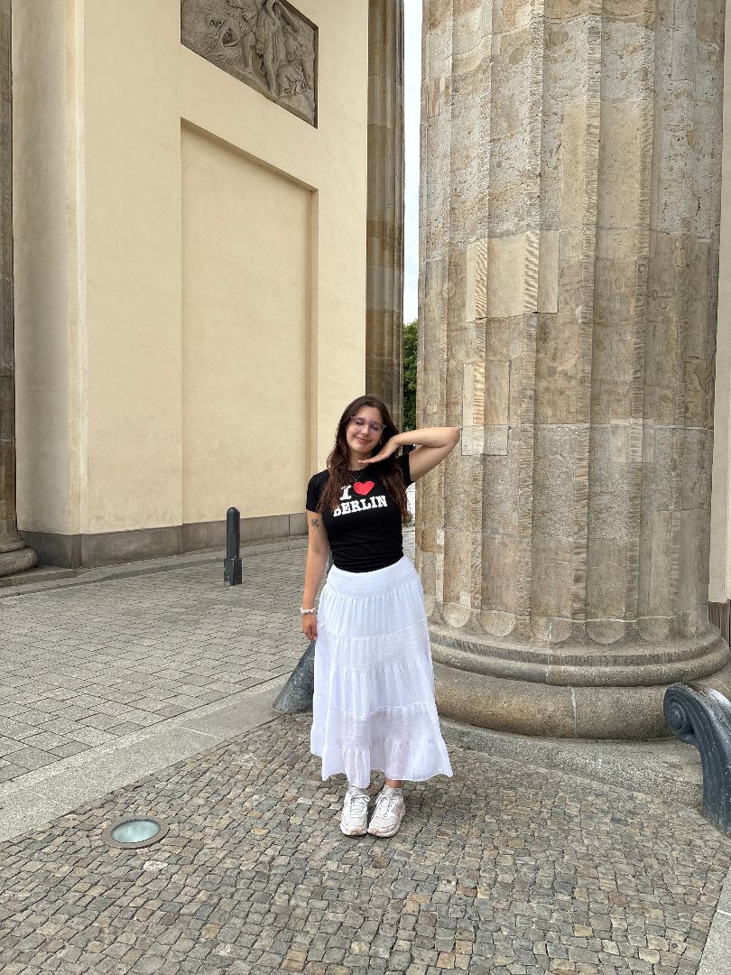 Alex wearing a white skirt and I Love Berlin shirt standing next to a large column in Germany